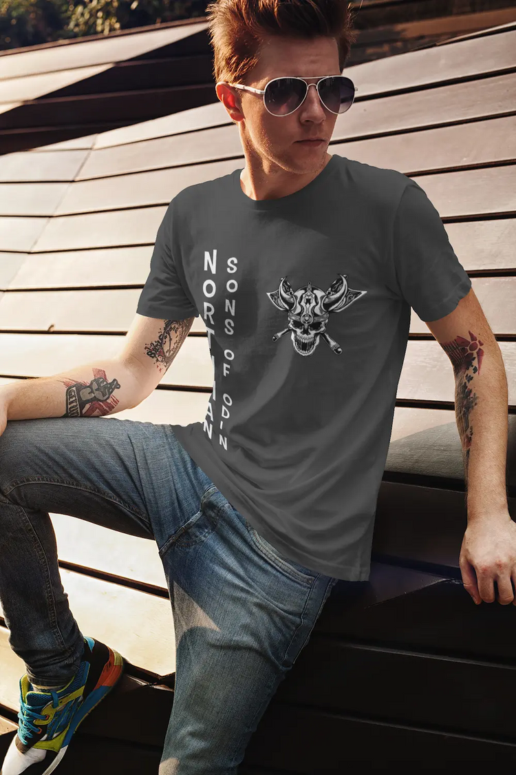 „Ein Mann mit Sonnenbrille sitzt lässig draußen in der Sonne und trägt ein graues T-Shirt der Northman & Sons of Odin Kollektion. Das handgroße weiße Wikinger-Skull-Wappen mit Kampfäxten ist auf der linken Brust zu sehen.“