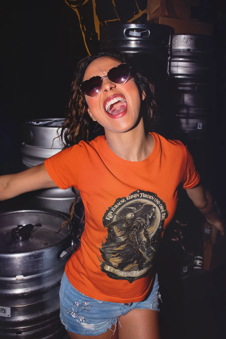 Frau im orangefarbenen Lonely Human Organic Shirt vor alten Metallfässern im Lager. Sie trägt eine Sonnenbrille. Streetstyle-Foto.