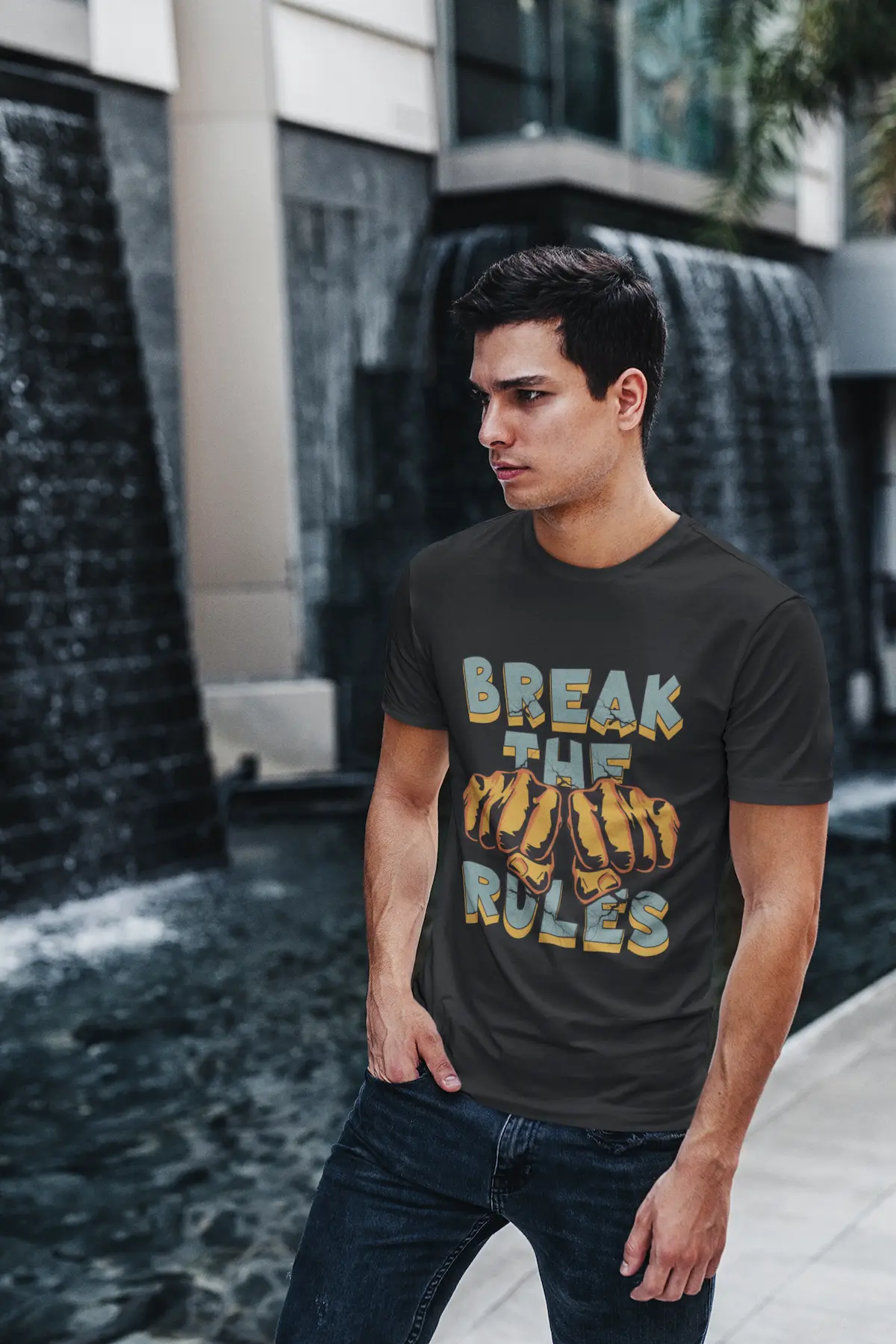 Lässiger Mann in schwarzem Organic Shirt und Jeans steht draußen. Auf dem Shirt ist das blaue BREAK THE RULES Motiv mit zerrissener Schrift und Fäusten zu sehen.