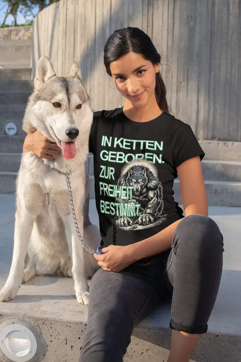 Frau in schwarzer Hose und schwarzem Bio Shirt sitzt und hält wolfsähnlichen Hund an einer Kette fest. Wolf Warrior Loyalität Motiv auf Organic Shirt.